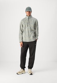 Fleece trui met groen geometrisch patroon, hoge kraag en halve rits. Zwarte taps toelopende broek en beige sport schoenen maken de outfit compleet.