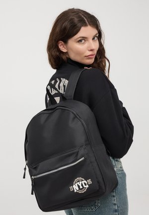 Zaino in nylon nero con tasca frontale con cerniera, dotato di un logo circolare "NYC 1995" e spallacci regolabili imbottiti.