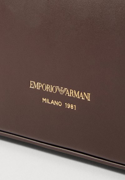 Smeđa kožna površina s upečatljivim zlatnim otisnutim tekstom "EMPORIO ARMANI MILANO 1981" s glatkom teksturom i čistim dizajnom.