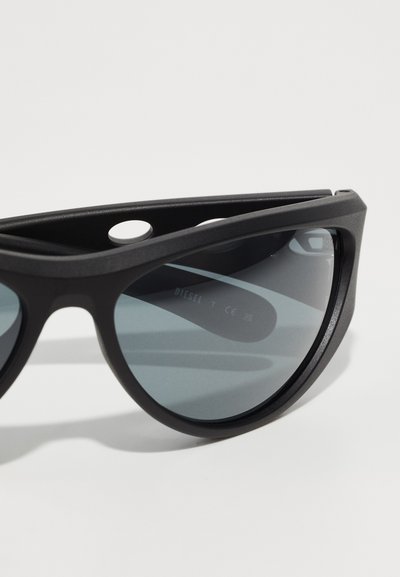 Lunettes de soleil noires avec une finition mate, verres ronds et un design de pont distinct comportant des découpes circulaires doubles. Logo visible sur le verre.