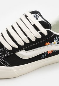 Sorte semskede joggesko med kremfargede lisser, med blomsterbroderi i oransje og blå på siden, og en hvit gummisåle.