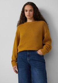 Kardigan w kolorze musztardowym, dzianinowy, z szerokim okrągłym dekoltem i ściągaczami, w połączeniu z ciemnoniebieskimi spodniami w stylu wide-leg. Widoczna teksturowana tkanina.