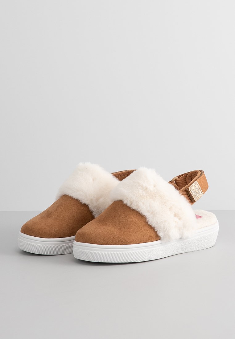 Zapatos tipo slip-on de ante marrón con una suave banda blanca y esponjosa, suela de goma y correa ajustable, que presenta un sutil brillo en el acento de la correa.