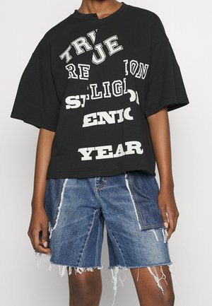 Persoon draagt een zwart oversized T-shirt met verspreide witte tekst en versleten patchwork denim shorts tegen een effen achtergrond.
