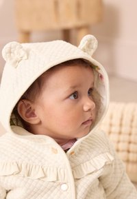 Veste de bébé ivoire matelassée avec des oreilles d'ours, un devant à volants et des boutons dorés. Doublée de tissu fleuri, avec une finition texturée.