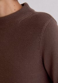 Brązowy sweter robiony na drutach z teksturowanym splotem, wysokim kołnierzem i widocznymi przeszyciami na ramionach.