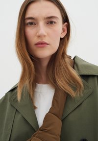 Trench coat verde oliva con un accento marrone sulla spalla, caratterizzato da un ampio colletto e dettagli con bottoni. Texture liscia, design minimalista.