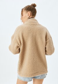 Beige, plüschiger Fleece-Jacke mit hohem Kragen, oversized Schnitt und strukturierter Oberfläche. Hat abgesetzte Schultern und Seitenschlitze am Saum.