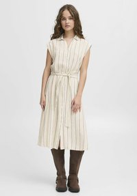 Cremefarbenes Midi-Kleid mit blauen vertikalen Streifen, kurzen Ärmeln, Knopfleiste und gebundenem Taillengürtel. Getragen mit braunen kniehohen Stiefeln.