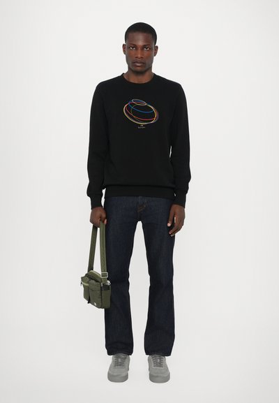 Schwarzer Pullover mit buntem, konzentrischem Kreisdesign, kombiniert mit dunklen Jeans. Das Model hält eine kleine olivgrüne Tasche mit einem Schulterriemen. Graue Sneakers.