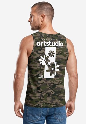 Muskelbepackter Mann trägt grünes Camouflage-Tanktop mit weißem Blumenmotiv und dem Text "artstudio" auf dem Rücken, kombiniert mit blauen Jeans.