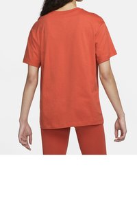 Kvinna klädd i en enkel, bränd orange t-shirt med kort ärm och matchande leggings, sedd bakifrån, stående mot en neutral bakgrund.