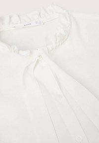 Camicia bianca con colletto arricciato e chiusura a bottoni, legata con un fiocco di tessuto a livello del collo.