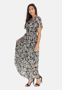 Robe maxi noire à fleurs avec des manches courtes, dotée d'une taille cintrée et d'un tissu fluide et transparent orné de fleurs blanches et jaunes.