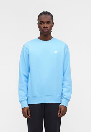Jeune homme aux cheveux tressés, portant un sweat-shirt Nike bleu clair et un pantalon noir, debout devant un fond blanc uni.