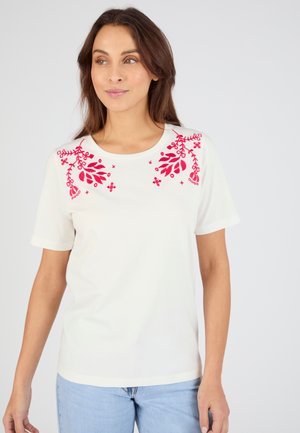 Femme aux cheveux bruns portant un haut blanc à manches courtes avec une broderie florale rouge sur les épaules, associé à un jean bleu clair, sur fond blanc.