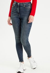 Jeansy skinny z wysokim stanem w niebieskim odcieniu z efektem sprania, posiadające pięć kieszeni i zamek błyskawiczny. W połączeniu z białymi butami sportowymi.