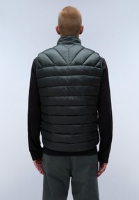 Mörkgrön vadderad väst med horisontella quiltade sektioner, hög krage och dragkedjeslutning, bärs över en svart långärmad skjorta.