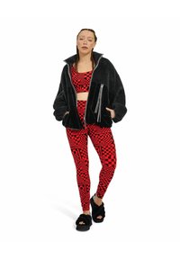 Giacca in fleece nera con zip, top corto a maniche corte con motivo rosso e leggings rossi con grafiche. Le ciabatte nere pelose completano l'outfit.