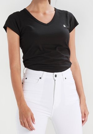 Femme portant un t-shirt noir ajusté à col en V et manches courtes, ainsi qu’un jean blanc taille haute avec des détails de boutons et de poches.