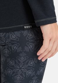 Czarny top z długim rękawem z metką Roxy, zestawiony z czarnymi legginsami w szary wzór płatków śniegu i gładką teksturą materiału.