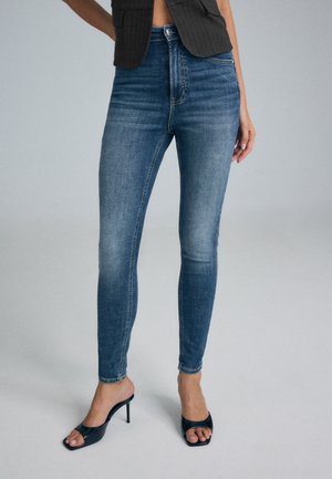 Un mannequin porte un jean bleu ajusté, un haut noir sans manches et des sandales noires à talons hauts sur un fond de studio uni.