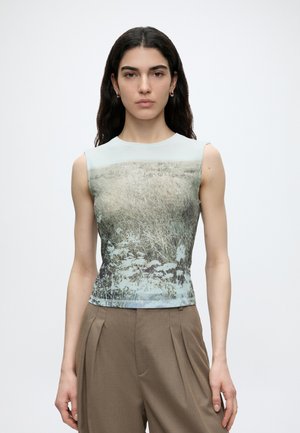 Jeune femme aux longs cheveux foncés portant un haut sans manches avec un imprimé de champ herbeux et un pantalon plissé marron taille haute, regardant vers l'avant.