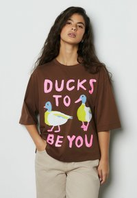 Tricou maro supradimensionat cu două rațe colorate și textul "DUCKS TO BE YOU" în roz. Mâneci scurte și decolteu rotund.