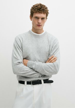Ung mand med krøllet hår iført en lysegrå sweater og hvide bukser med et sort flettet bælte, stående med armene over kors.