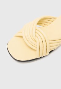 Gul imitativ lädersandal med vriden remdesign, fyrkantig tå och svart platt sula. Har en slät yta och minimalistisk stil.