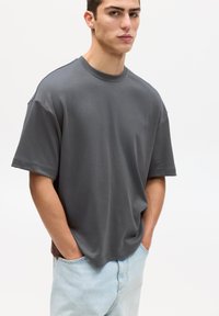 T-shirt gris oversized avec un col rond et des manches courtes, fabriqué à partir d'un tissu doux ; le mannequin porte un jean bleu clair avec des poches.