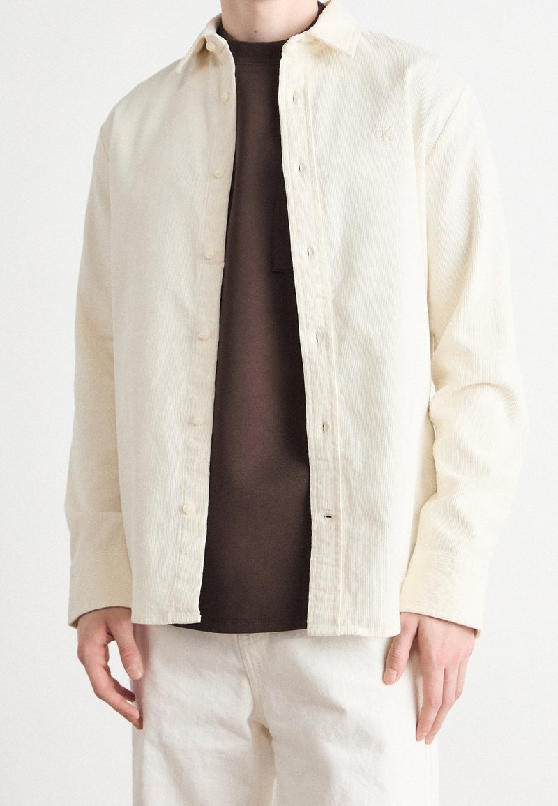 Camicia in velluto a coste color crema con chiusura a bottoni, caratterizzata da una leggera texture e un logo sul petto, indossata sopra una camicia marrone e pantaloni chiari.