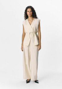 Top beige senza maniche con dettaglio a legatura, abbinato a pantaloni a gamba larga. Il tessuto sembra testurizzato, complementato da scarpe nere a punta.