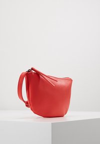 Červená, texturovaná kožená taška se zipovým uzávěrem a zápěstním popruhem. Zaoblený tvar s minimalistickým designem a akcentovaným prošíváním.