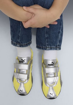 Kind draagt gele en zilveren Puma sneakers met klittenbandsluitingen, witte sokken en blauwe spijkerbroek, handen gevouwen boven de knieën.