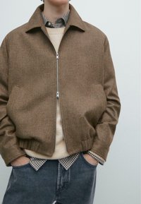 Bruine jas van wolmix met rits en een gestructureerde silhouet, zijzakken, gedragen over een beige gebreide trui en een geruit overhemd.