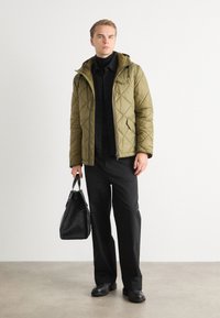 Veste matelassée vert olive avec capuche, fermeture éclair, portée sur une chemise et un pantalon noirs. Tenant un sac noir texturé, debout sur un sol gris.