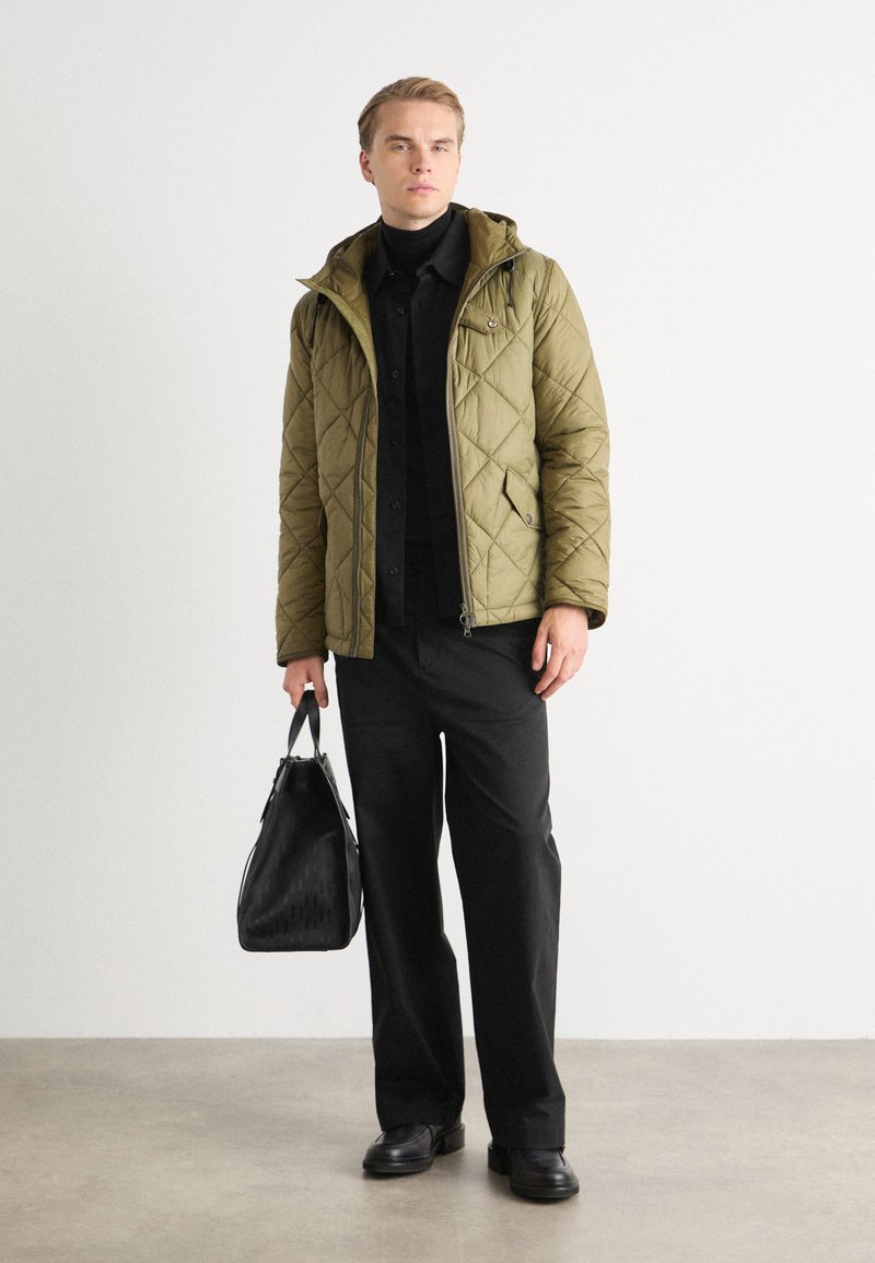 Giacca trapuntata verde oliva con cappuccio, chiusura con zip, indossata sopra una camicia nera e pantaloni. Tiene una borsa nera con texture, in piedi su un pavimento grigio.