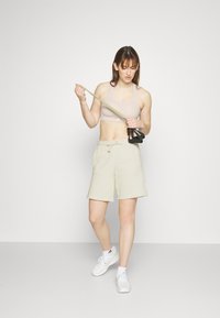 Shorts deportivos de color beige claro hechos de una tela suave, combinados con un sujetador deportivo a juego. La figura sostiene una banda de resistencia y un mango negro.