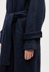 Cappotto lungo in lana blu scuro con finitura testurizzata, vita cintrata da una cintura e maniche con risvolto che presentano un dettaglio a bottoni. Primo piano del braccio e della mano.