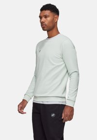 Felpa verde menta con collo tondo, polsini a coste e fondo a coste. Piccolo dettaglio ricamato sul petto. Indossata sopra una camicia bianca e abbinata a pantaloni neri.