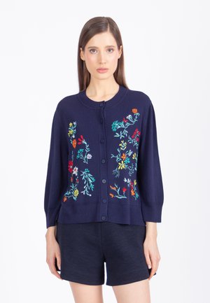 Žena oblečená v námornícky modrom kardigane na gombíky s farebnou kvetinovou výšivkou, v kombinácii s námornícky modrými šortkami, stojaca na bielom pozadí.