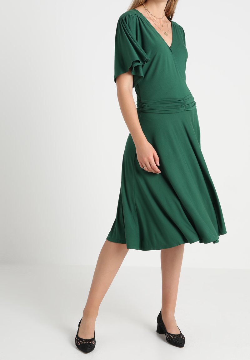 Femme portant une robe verte longueur genou avec manches flottantes et taille froncée, associée à des chaussures noires à talons bas, debout sur un fond blanc.