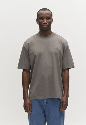 Mees seisab näoga ettepoole, seljas liibuv hall T-särk ja sinised teksad, taust on hele ja lihtne.