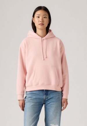 Pink hooded sweatshirt in soft fabric with a kangaroo pocket, drawstring hood, and ribbed cuffs, paired with light blue distressed jeans.