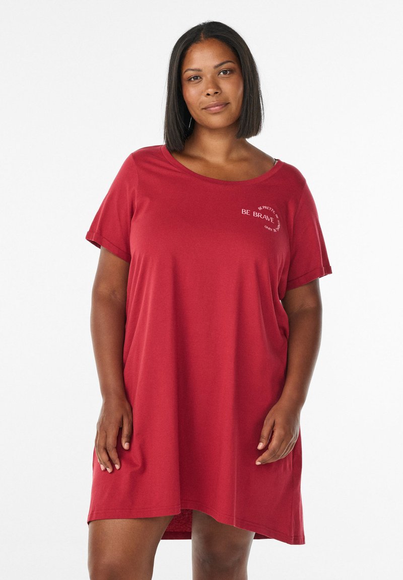 Woman standing, wearing a loose red knee-length dress with "Be Brave" text near the neckline, against a plain white background.