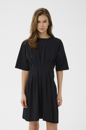 Mujer con cabello castaño ondulado que lleva un vestido negro de manga corta, plisado y hasta la rodilla, de pie contra un fondo claro y liso.