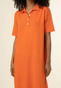 Femme portant une robe polo orange vif, à manches courtes et longueur au genou, avec une patte à trois boutons et un col, sur un fond uni.