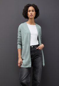 Světle zelený cardigan z texturovaného materiálu, s otevřeným předním designem, dlouhými rukávy, v kombinaci s bílým topem a tmavými džíny.