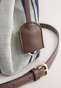 Sac en toile avec rayures bleu marine et blanches, équipé d'une sangle en cuir marron et d'un patch en cuir arrondi avec anneau métallique.